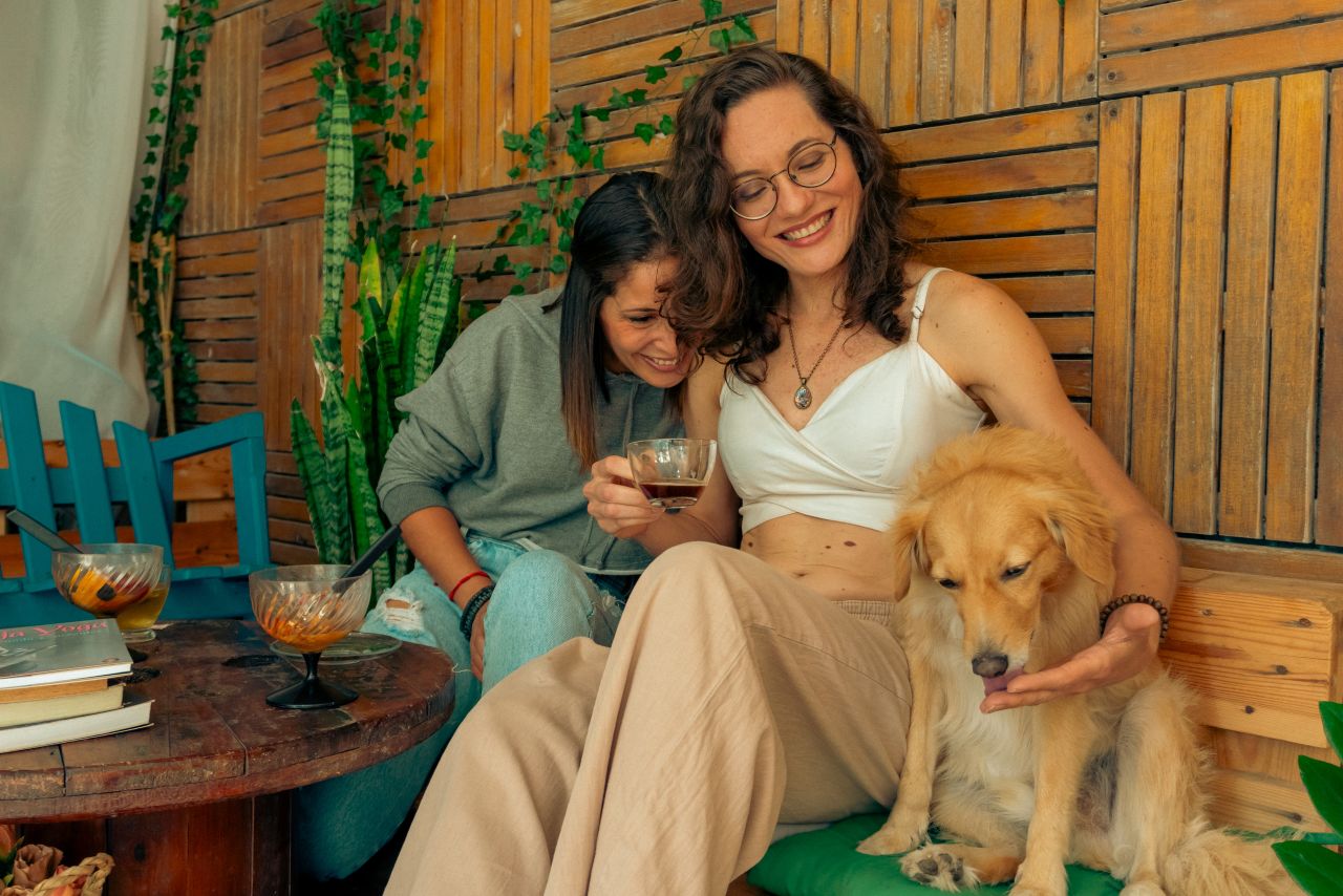 Two women enjoying drinks and laughter with a dog in a cozy indoor setting.