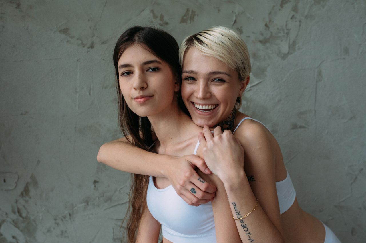 About Two young women smiling and hugging against a textured concrete wall, expressing friendship and joy.